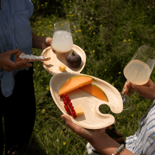 Zwei Personen halten die Cocktailplates aus Palmblatt mit Snacks und Obst sowie ein Weinglas bei einer Feier.