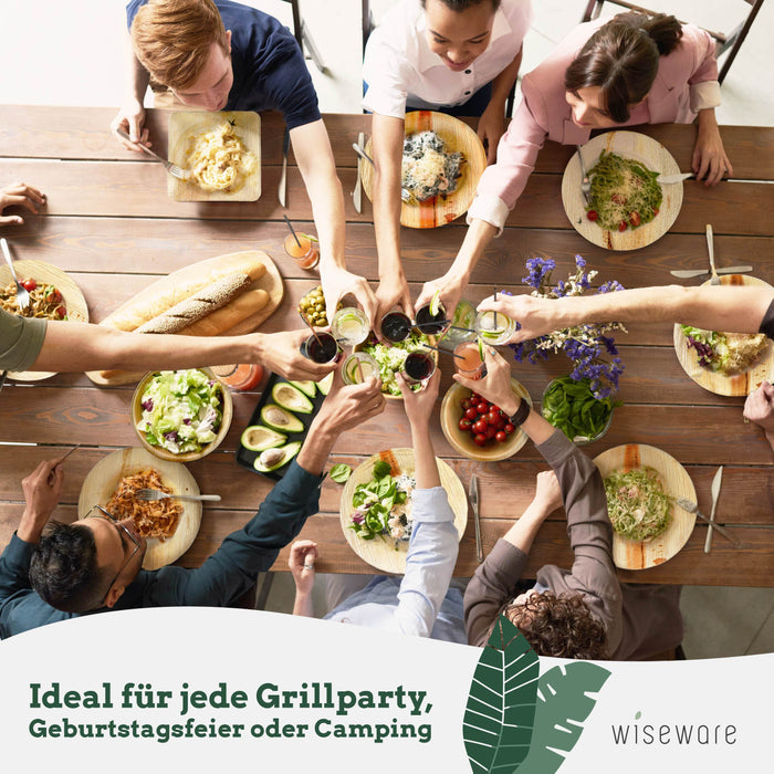 Eine Gruppe stößt mit ihren Gläsern gemeinsam über einem Tisch an, verschiedene Pastagerichte und Beilagen sind auf Palmblatttellern und Palmblattschalen serviert.